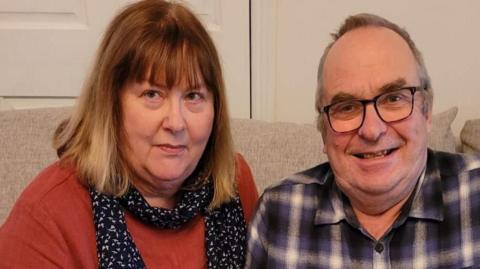 Mags and Dewi sit together. Mags is wearing a red jumper and black and white scarf. She has short blond/brown hair. Dewi is grinning, he has grey hair and is wearing glasses. 