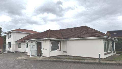 A white rendered single storey building with a brown tiled roof. A white door is open at the front with a path leading to a brick paved road.