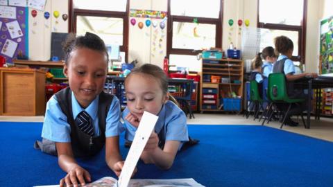 Two girls in a classroom looking at a book together. They are next to each other on a mat looking at the book and wearing school uniform. In the back right of the image there are three more children sitting on chairs with their backs to the camera, along the walls are various bits of shelving and drawers