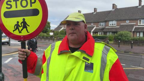 Martin is wearing a bright flourescent yellow coat and hat. He is holding a yellow and red school crossing sign (lollipop)