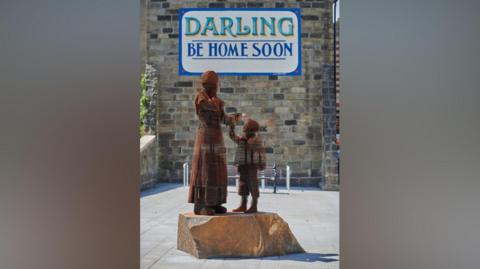 The sculpture of a mother and son stands in front of a sign reads "Darling Be Home Soon".