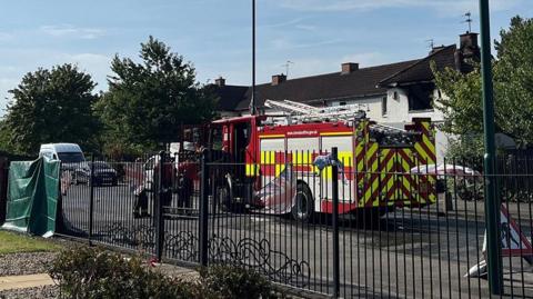 A fire engine has parked outside residential homes in Middlesbrough. Fire fighters are leaving the vehicle. A black fence is lining the near side of the street before the white houses on the opposite side.