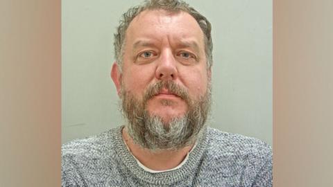 Mugshot of Matthew Gilkes. He is wearing a grey marled sweater and has greying short curly hair and a beard.