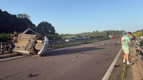 A silver car is pictured on its roof and up against the central reservation of the motorway. A man in a blue t-shirt and beige shorts is standing on the side of the motorway on the phone. Several other people are pictured in the background standing on the motorway. Debris is scattered across the road.