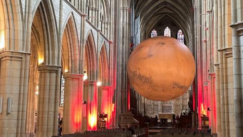 The Mars sculpture. It is an orange and brown globe hanging from the ceiling of Truro Cathedral. Either side are supporting columns of the cathedral and stained glass windows behind it.