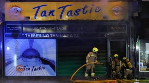 fire crews stand in the dark with a fire house outside the burnt-out Tan Tastic salon