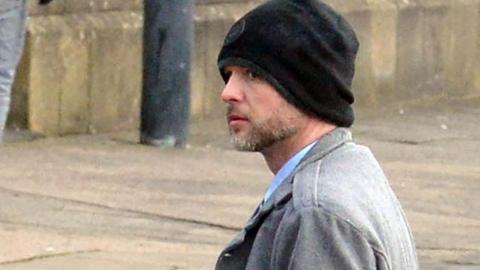 William Coull leaving court in Glasgow. He is standing on the street wearing a black beanie hat and a grey jacket over a light blue shirt, the collar of which can just be seen. He has a short greying beard. He is standing in front of two yellow lines on the road.