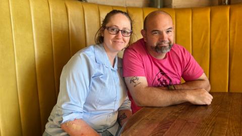 Amy and James sit on a yellow sofa in front of a wooden table. Amy is on the left and has a light blue shirt on and is wearing glasses with a purple frame. Her brown hair is tied back and she's leaning towards James. He has a pink t-shirt on and has a shaved beard. His arms are crossed on the table.