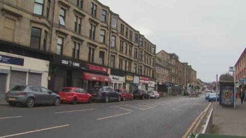 Neilston Road, Paisley,  street with cars parked on either side, image taken from the pavement near a bus stop with tenement flats opposite and shops on the ground floor