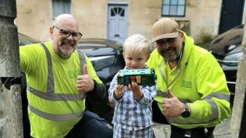 Two bin collectors in high visibility clothing smile and hold their thumbs up to the camera, as a young boy stands between them holding a toy recycling truck. The boy wears blue and white checked pyjamas. One of the bin collectors wears a brown cap, while the other has glasses on and a bald head. 