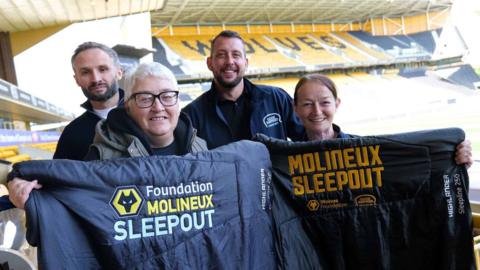 Four people are standing inside a football stadium. Two men are standing at the back, wearing jackets that read "Good Shepherd". Two women are in front holding sleeping bags that read "Molineux sleepout". There are thousands of football seats in stands behind them in a gold colour. The word "Wolves" is spelt out in black on them. 