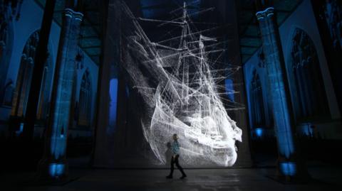 A light show inside a large minster church. A woman walks past an illuminated installation of a huge white sailing ship. The walls of the minster are lit up in blue, bringing out details of the medieval architecture.