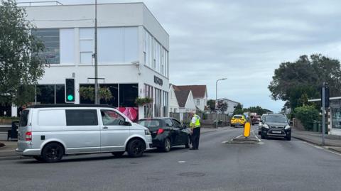 Emergency services are at the scene of a two‑vehicle road traffic collision in Jersey. A police officer is seen directing traffic away from the road closure. 