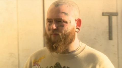 Aaron Connolly. He is bald with light brown beard, wearing a yellow t-shirt. He has tattoos on his head and face and an eyebrow piercing.