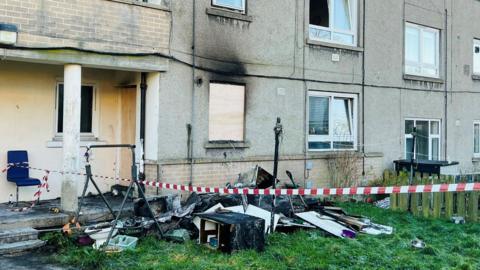 Image of flats after fire, charred walls and damaged items on grass, with red and white taping.