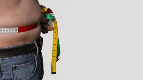 A man holding a tape measure around his bare tummy. He is wearing jeans.