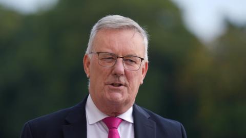 Mike Nesbitt is wearing a white shirt and pink tie. He has a navy blazer on. He is also wearing black glasses. The background behind him is blurry. 