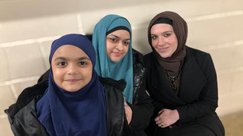 Nisha wearing a blue scarf, Moneera, a young girl in a blue scarf and black coat and Eva, wearing a brown scarf, smiling for the camera.