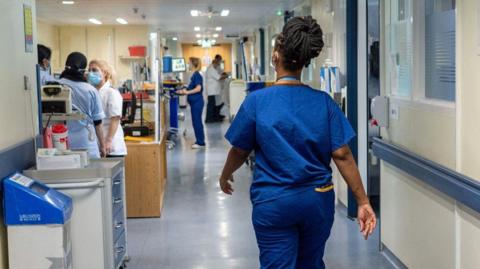 A woman wearing a blue NHS staff uniform walks through the corridor of a hospital away from us.