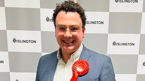 Oliver Steadman, wearing a red "Vote Labour" rosette" and standing in front of a backdrop reading "Islington".