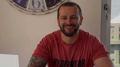 A man in a red T-shirt with short dark hair and a beard.