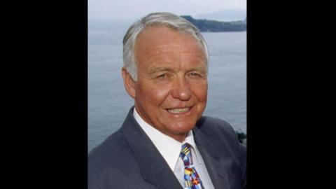 Craig Rich seen wearing a navy suit, white shirt and patterned tie. He is pictured with the sea in the backhround.