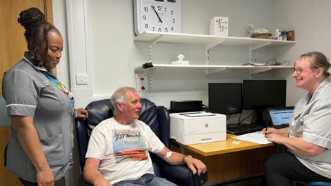 Ian Robinson sits on a chair in a consulting room. Sitting at a desk to his right is a female member of medical staff who is smiling at him as he speaks. Another female member of medical staff is standing to his left with her hand resting on the back of his chair. 