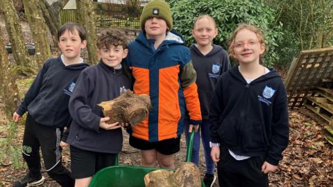 Five children dressed in school uniform in a wooded area using a wheelbarrow to carry wood 