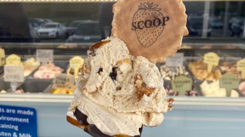 A hand holds an ice cream cone with two scoops, chocolate coating, caramel drizzle and a biscuit on top, with a display of ice cream tubs in the background.