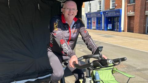 A bald man with glasses wearing black lycra with red and white lettering, sitting on a stationary bike  on a street with his hands on the handlbars and smiling at the camera.