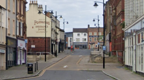 A one-way street with buildings to the left, right, and straight ahead. One of the buildings has a sign on the side saying 'Potteries Insurance Brokers'