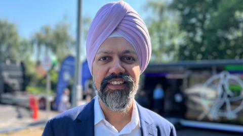 Slough MP Tan Dhesi is wearing a pink turban as well as a white shirt and navy suit. He is smiling and the pop-up heart clinic is visible in the background. 