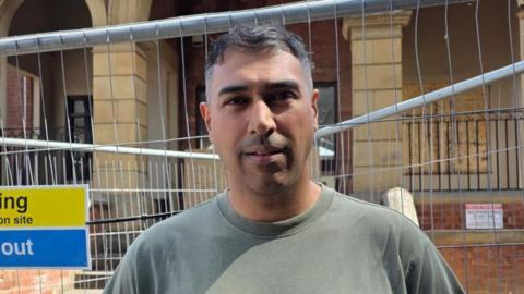 A man in a green t-shirt stands in front of railings next to a large old brick building 