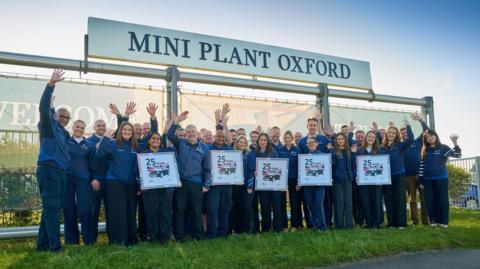 A group of workers standing underneath the "Mini Plant Oxford" sign. They are all wearing blue and some are holding signs marking the anniversary.