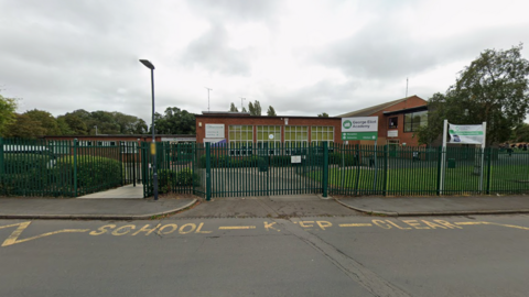 Green railings and gates near a pavement by a road. The street has the words school keep clear in yellow on the ground. Behind the railings is a sign saying George Eliot Academy and a building is in the background behind it.