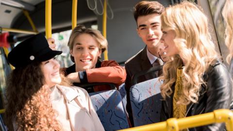 A woman with long brown hair and a light coat wearing a dark hat, A woman with long blonde hair with a black jacket. A man with blonde hair and an orange and blue jacket and a man with brown hair, white shirt, dark blazer and a dark tie. They are all on a bus and smiling.