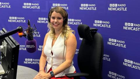 Kim McGuinness sitting in front of a microphone in the BBC Radio Newcastle studio wearing headphones. The BBC Newcastle banner is behind her. She has a pair of headphones over her long light brown hair and is wearing a white short-sleeved top.