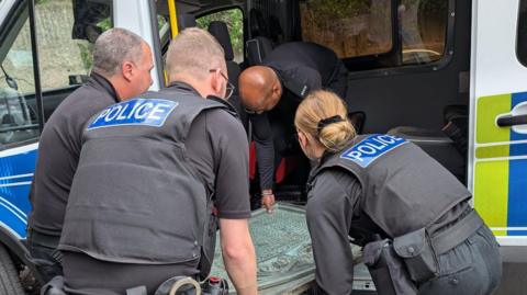 Four people in black police uniforms lifting a memorial plaque out of the side of a police van. The plaque is large and appears blue with age.