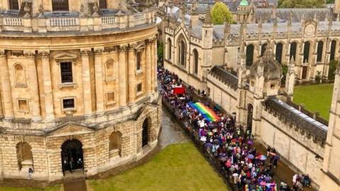 A colourful parade including a large rainbow banner passes Oxford's Radcliffe Camera