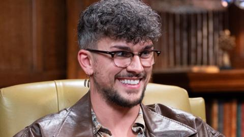 Jack from Traitors is smiling while sat in a library and wearing a brown leather jacket