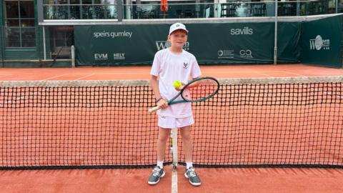 A young boy in tennis whites holding a racket and ball on a tennis court.