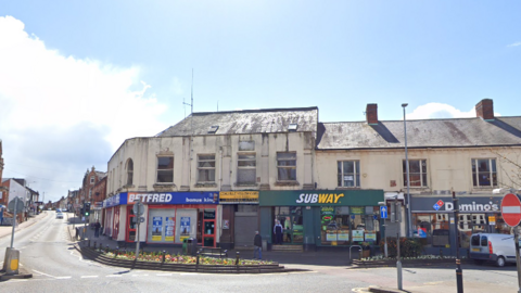 A Google Streetview image of the Marlborough Centre which is a two-storey building in poor condition. It has shopfronts on the groundfloor including a Betfred which is red and blue, and a Subway which is green.