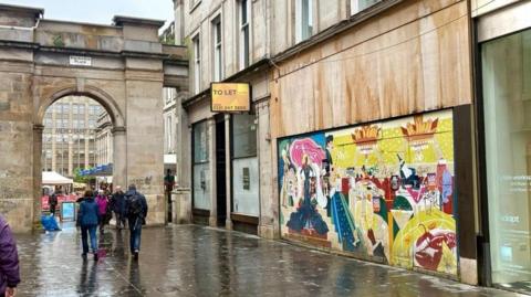 Wide street view of Exchange Place in Glasgow showing a colourful mural covering the boarded frontage of Rogano restaurant, with pedestrians walking past and a stone archway marked Merchant City in the background.