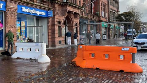The entrance to the pedestrian area by the Crown and Mitre in Carlisle, which is a large and beautiful redbrick building with leaded windows, a first floor balcony running most of its length, ornate stone work and many chimneys. The ground floor is given over to shop fronts. A taxi is parked behind a large, plastic temporary orange barrier, next to which is a white barrier of the same kind just outside a toy shop.