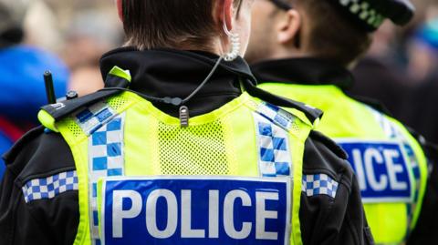 Two police officers wearing yellow hi-vis vests with "police" written on the back. One officer has an earpiece. They both have their backs to the camera.