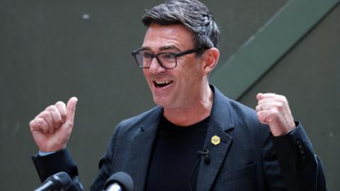 Andy Burnham, wearing a black jacket and t-shirt, stands at a podium.