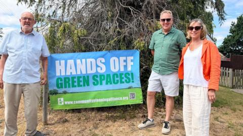 A sign reads 'hands off green spaces' in front of a tree and set on browning grass. On the left is a man in cream chinos and a blue shirt, while a man with white shorts and a green shirt, and a woman in cream trousers and an orange jacket stands on the right,.