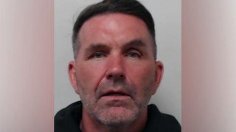 A mugshot of a man with short dark hair wearing a dark top.
