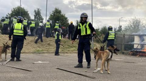 Police officers in riot gear stand in a line. Two of the officers are holding dogs on leads.