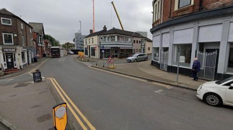A street view image of a crossroads junction in a town centre. Shops and other high street premises are either side of the road. A white car is parked on the left, a lorry in the distance and a silver car ahead and to the right. 